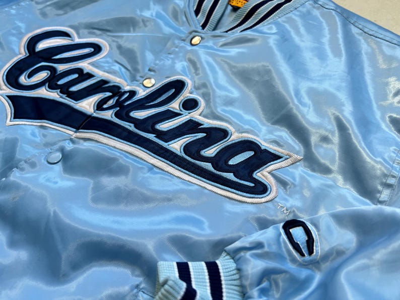 K&ouml;nnte beinhalten: Hellblaue Satinjacke mit dem Wort "Carolina" in Marineblau und Wei&szlig; gestickt. Die Jacke hat einen gerippten Kragen und Manschetten mit marineblauen und wei&szlig;en Streifen. Die Jacke hat einen Druckknopfverschluss.