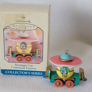 May include: A Hallmark Keepsake Ornament, a pastel-colored passenger car from the Cottontail Express train set. The car is decorated with a blue bird and has a pink roof with a blue dome on top. The ornament is in its original box, which features the Hallmark logo and the text "Passenger Car Cottontail Express Collector's Series."
