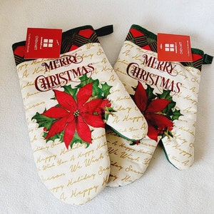 Pode incluir: Um par de luvas de forno de cor creme com um design de poinsettia vermelha e o texto "Merry Christmas" em escrita dourada. As luvas são feitas de algodão e têm uma textura acolchoada.