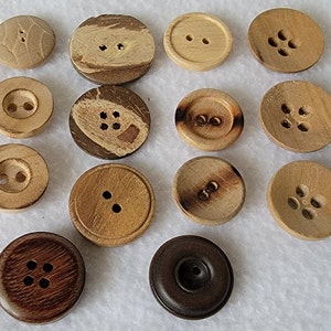 May include: Assortment of 14 wooden buttons in various shapes, sizes, and colors. Some buttons have a natural wood finish, while others have a dark brown stain. The buttons have different numbers of holes, ranging from two to five.