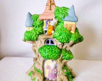 May include: A ceramic Easter tree house with a pink roof and a cross on top. The tree house has a door and windows, and there are three bunnies on the tree. The tree is decorated with green leaves and flowers.