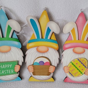 May include: Three wooden Easter gnomes with bunny ears. The gnome on the left is holding a sign that says "Happy Easter". The gnome in the middle is holding a basket with Easter eggs. The gnome on the right is holding a decorated Easter egg.