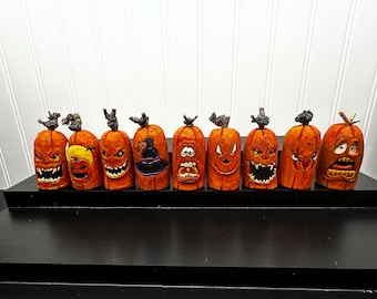 May include: A collection of eight carved wooden pumpkins with various facial expressions. Each pumpkin is orange with a dark brown stem. Some pumpkins have carved teeth and angry faces, while others have surprised or silly expressions. The pumpkins are displayed on a black surface.