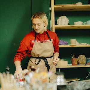 Puede incluir: Una persona con una sudadera roja con el texto "MAKE BETTER CHOICES" y un delantal color beige en un estudio de cerámica. Estantes exhiben cerámica y materiales de arte. Una lámpara blanca cuelga del techo.