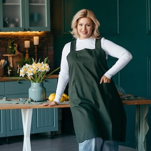 May include: A woman wearing a dark green apron with a white shirt and jeans. She is standing in a kitchen with a table in front of her. There are lemons on the table.
