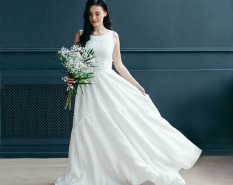 May include: A sleeveless white dress with a flowing skirt. The dress is made of a light fabric and has a simple, elegant design. The person is holding a bouquet of white flowers and greenery.