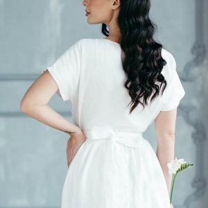 A white linen dress with a tie waist and short sleeves. The dress is flowing and has a relaxed fit. The model is holding a single white flower.