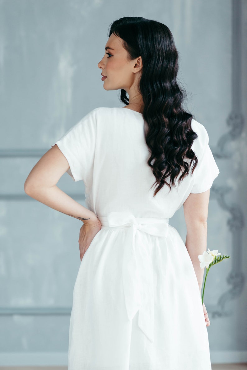 A white linen dress with a tie waist and short sleeves. The dress is flowing and has a relaxed fit. The model is holding a single white flower.
