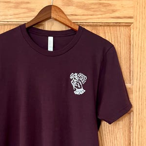 Puede incluir: Camiseta de color granate con un gráfico blanco en el pecho izquierdo. El gráfico muestra una mano sosteniendo un ramo de flores con la inscripción "Good Luck". La camiseta está colgada en una percha de madera.