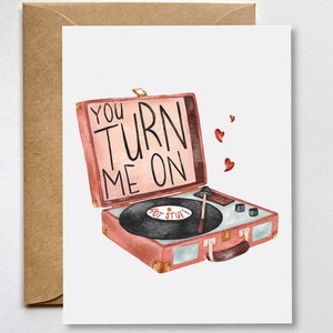 May include: A pink and white vintage-style record player with the text "You Turn Me On" on the lid. The record player has a black record on the turntable and a red needle. There are small red hearts floating above the record player.