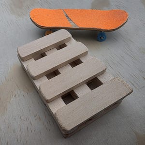 May include: A miniature skateboard with an orange deck and blue wheels rests above a small wooden ramp structure. The ramp is made of light-colored wood, with a slatted design. The background is a light wood surface.