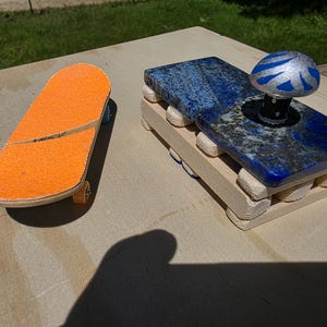 May include: An orange finger skateboard with a split design sits next to a blue and silver mushroom-shaped object on a wooden base. The mushroom is on a blue stone base. The skateboard has small wheels.