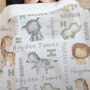 K&ouml;nnte beinhalten: Eine wei&szlig;e Babydecke mit einem sich wiederholenden Muster aus verschiedenen Tieren, darunter eine Giraffe, ein Zebra, ein Elefant, ein L&ouml;we und ein Nashorn. Die Decke tr&auml;gt den Namen "Hayden James" in hellrosa Schrift.