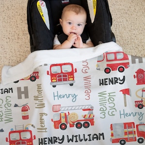 May include: A white baby blanket with a red fire truck pattern. The blanket features the name "Henry William" repeated throughout the design. The blanket is shown in a car seat with a baby sitting in it.