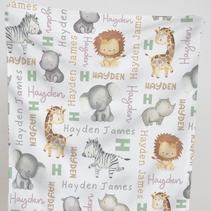 K&ouml;nnte beinhalten: Eine wei&szlig;e Babydecke mit einem sich wiederholenden Muster aus verschiedenen Dschungeltieren, darunter eine Giraffe, ein Zebra, ein L&ouml;we, ein Elefant und ein Nashorn. Die Decke ist mit den Namen "Hayden" und "James" in rosa und gr&uuml;ner Schrift verziert.