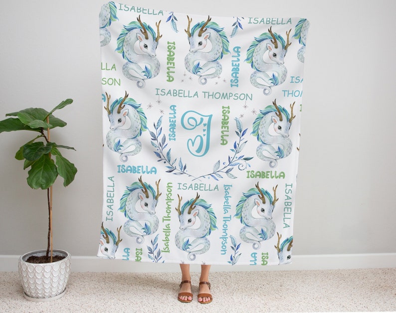 May include: A white baby blanket with a repeating pattern of blue and green watercolor dragons. The blanket features the name "Isabella" and the initial "J" in a light blue and green font.