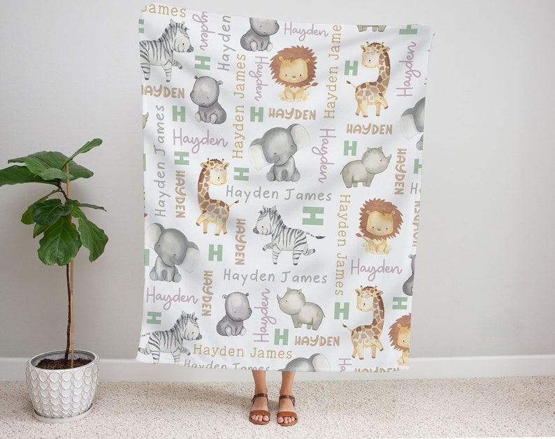 K&ouml;nnte beinhalten: Eine wei&szlig;e Babydecke mit einem sich wiederholenden Muster aus verschiedenen Dschungeltieren, darunter eine Giraffe, ein Zebra, ein L&ouml;we, ein Elefant und ein Nashorn. Die Decke ist mit den Namen "Hayden" und "James" in rosa und gr&uuml;ner Schrift verziert.