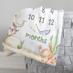 May include: A white baby milestone blanket with a blue, green and orange underwater theme. The blanket features an octopus, a stingray, a crab and seaweed. The numbers 8 through 12 are printed on the blanket, along with the word "months".
