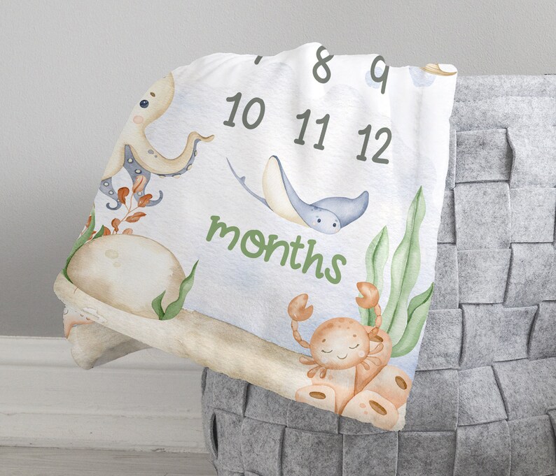 May include: A white baby milestone blanket with a blue, green and orange underwater theme. The blanket features an octopus, a stingray, a crab and seaweed. The numbers 8 through 12 are printed on the blanket, along with the word "months".