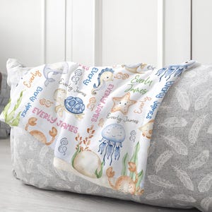 May include: A white baby blanket with a sea life theme. The blanket features a variety of sea creatures, including a seahorse, a starfish, a turtle, a crab, a jellyfish, and a dolphin. The blanket is decorated with the name "Everly James" repeated throughout.