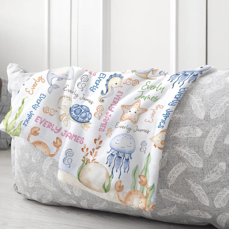 May include: A white baby blanket with a sea life theme. The blanket features a variety of sea creatures, including a seahorse, a starfish, a turtle, a crab, a jellyfish, and a dolphin. The blanket is decorated with the name "Everly James" repeated throughout.