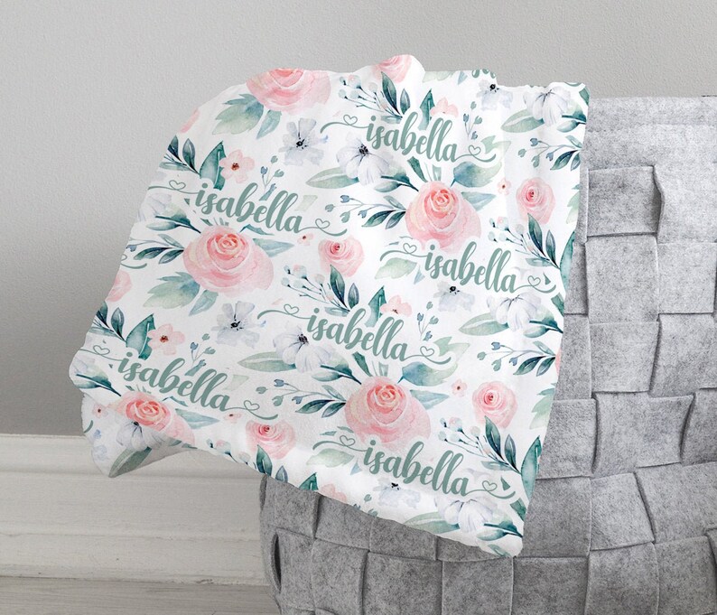 May include: A white baby blanket with a floral pattern featuring pink roses, green leaves, and white flowers. The name "isabella" is repeated throughout the design.