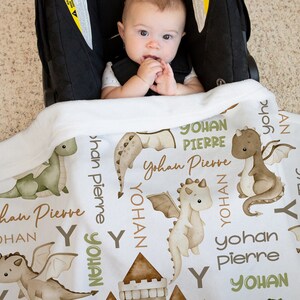 May include: White baby blanket with a green and brown dragon pattern. The blanket has the name "Yohan Pierre" repeated throughout the design.