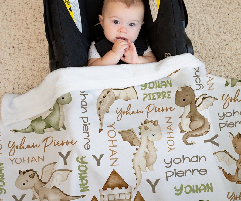 May include: White baby blanket with a green and brown dragon pattern. The blanket has the name "Yohan Pierre" repeated throughout the design.
