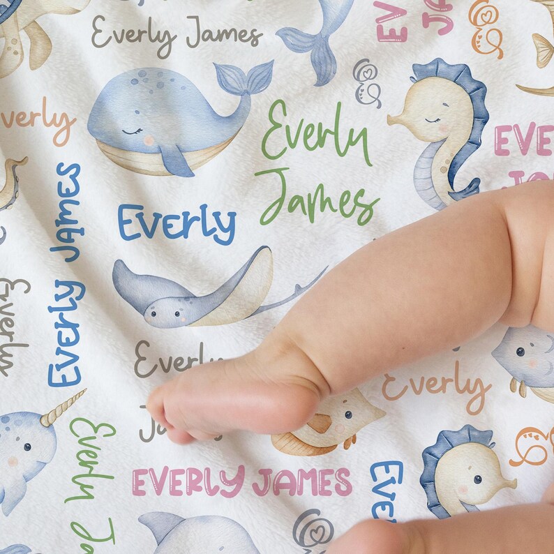 May include: A white baby blanket with a sea life theme. The blanket features a variety of sea creatures, including whales, seahorses, and stingrays. The blanket is decorated with the name "Everly James" repeated throughout.