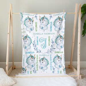 May include: A white baby blanket with a blue and green dragon design. The blanket features the name "Isabella Thompson" and the letter "I" in a floral wreath.