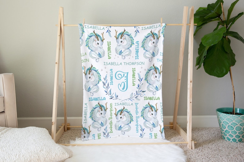 May include: A white baby blanket with a blue and green dragon design. The blanket features the name "Isabella Thompson" and the letter "I" in a floral wreath.
