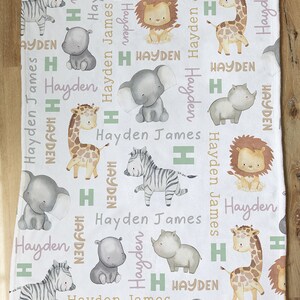 K&ouml;nnte beinhalten: Wei&szlig;e Babydecke mit einem sich wiederholenden Muster aus verschiedenen Tieren, darunter ein Zebra, eine Giraffe, ein Elefant, ein Nashorn, ein L&ouml;we und ein Nilpferd. Die Decke ist mit dem Namen Hayden James in rosa und gr&uuml;ner Schrift verziert.