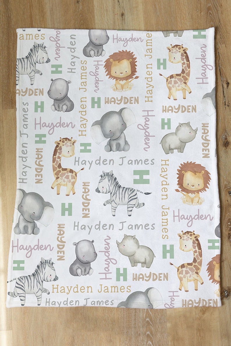 K&ouml;nnte beinhalten: Wei&szlig;e Babydecke mit einem sich wiederholenden Muster aus verschiedenen Tieren, darunter ein Zebra, eine Giraffe, ein Elefant, ein Nashorn, ein L&ouml;we und ein Nilpferd. Die Decke ist mit dem Namen Hayden James in rosa und gr&uuml;ner Schrift verziert.