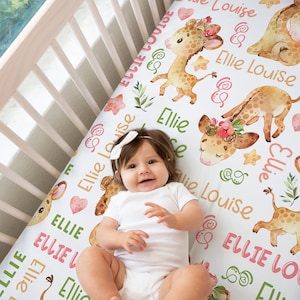 May include: White crib sheet with a repeating pattern of watercolor giraffes, green leaves, and the name "Ellie Louise" in pink and green lettering.