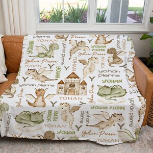 May include: White fleece blanket with a repeating pattern of green and brown dragons, a castle, and the names "Yohan" and "Pierre".
