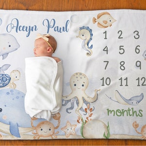 May include: A white baby blanket with a blue and green ocean theme. The blanket features a whale, octopus, seahorse, turtle, crab, and fish. The blanket also has a number chart for tracking a baby's growth. The text on the blanket reads "Aceyn Paul" and "months".