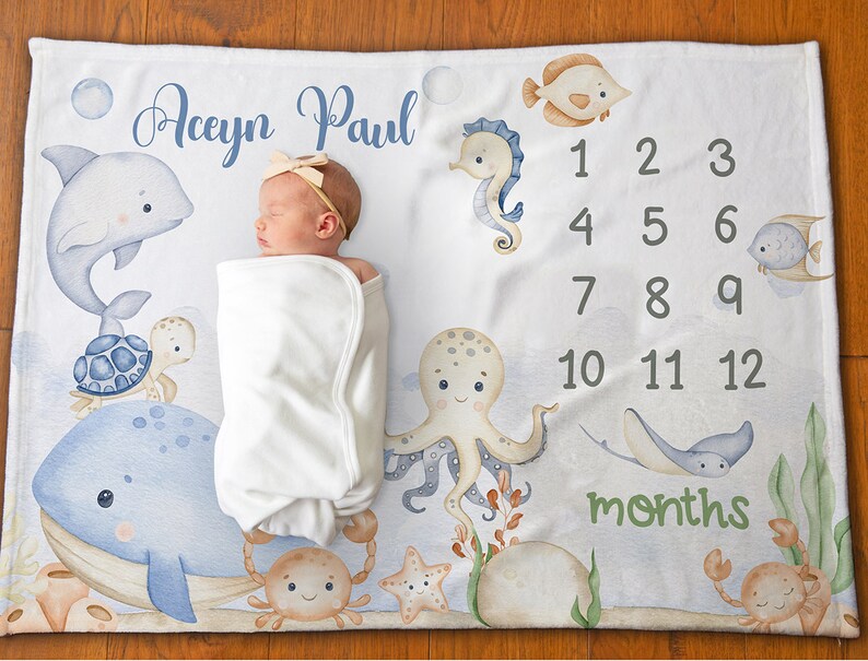 May include: A white baby blanket with a blue and green ocean theme. The blanket features a whale, octopus, seahorse, turtle, crab, and fish. The blanket also has a number chart for tracking a baby's growth. The text on the blanket reads "Aceyn Paul" and "months".