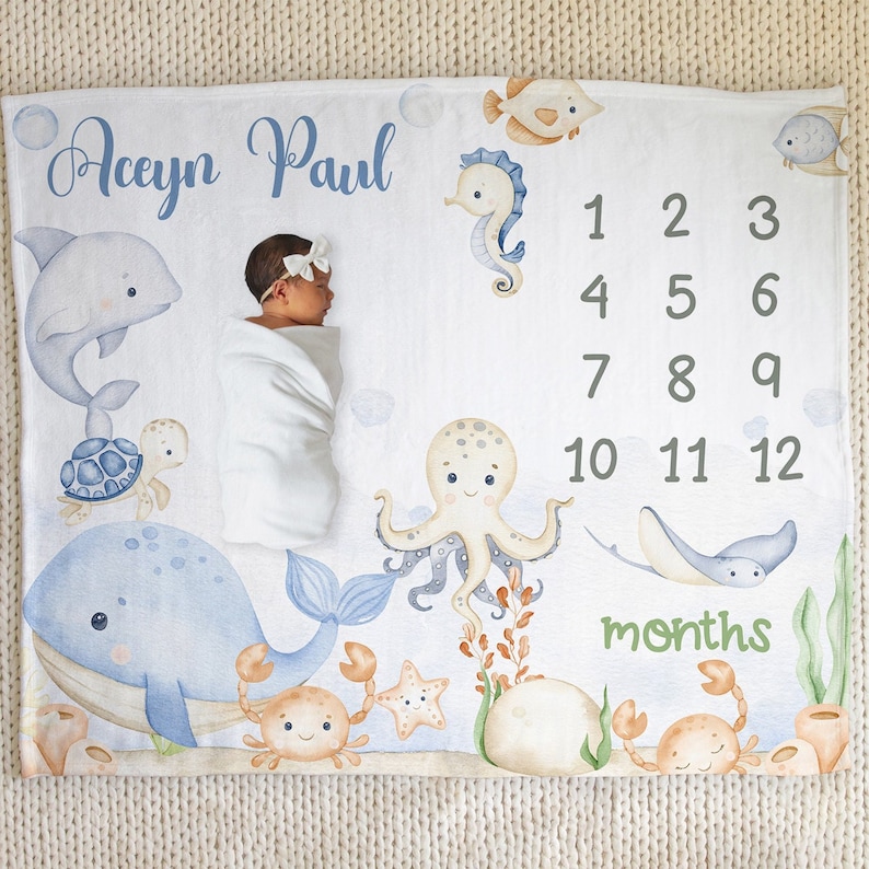 May include: A white baby blanket with a blue and white ocean theme. The blanket features a whale, octopus, seahorse, starfish, crab, and turtle. The blanket also has a number chart for tracking a baby's growth in months. The text "Aceyn Paul" is written at the top of the blanket.