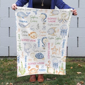 May include: A white baby blanket with a sea-themed pattern. The blanket features various sea creatures, including seahorses, jellyfish, starfish, and a turtle. The name "Sophie Laine" is repeated throughout the design in pink, blue, and green lettering.
