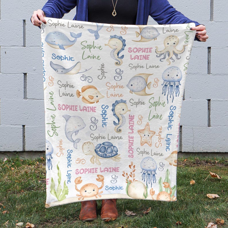 May include: A white baby blanket with a sea-themed pattern. The blanket features various sea creatures, including seahorses, jellyfish, starfish, and a turtle. The name "Sophie Laine" is repeated throughout the design in pink, blue, and green lettering.