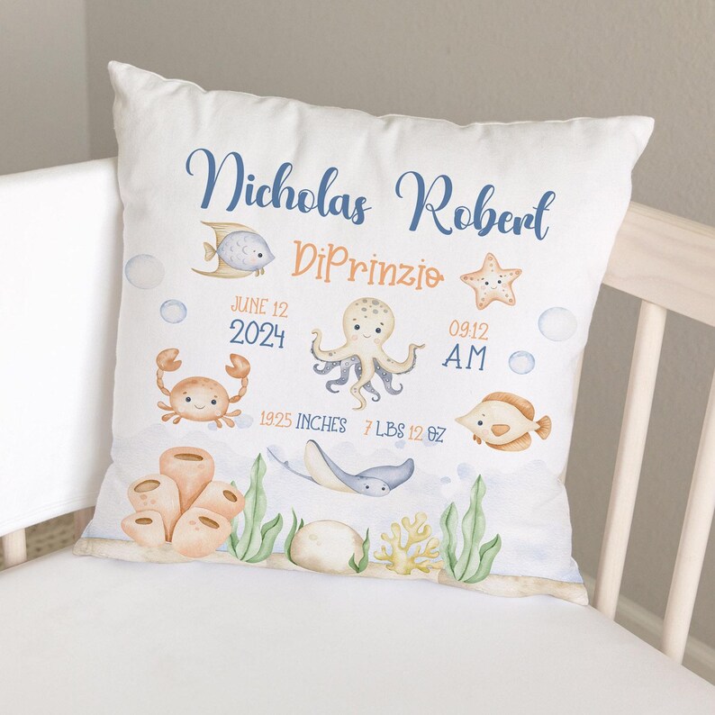 May include: A white pillow with a blue and orange ocean-themed design. It features a starfish, an octopus, a crab, a fish, and a stingray. The text on the pillow reads "Nicholas Robert DiPrinzio June 12, 2024 09:12 AM 19.25 inches 7 lbs 12 oz."