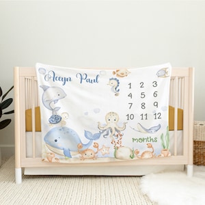 May include: A white baby blanket with a blue and white ocean theme. It features a whale, octopus, seahorse, turtle, and crab. The blanket has a personalised name, "Aceyin Paul", and a number chart for tracking a baby's growth in months.