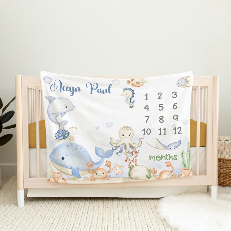 May include: A white baby blanket with a blue and white ocean theme. It features a whale, octopus, seahorse, turtle, and crab. The blanket has a personalised name, "Aceyin Paul", and a number chart for tracking a baby's growth in months.
