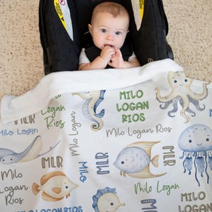 May include: A white baby blanket with a sea life theme. The blanket features a variety of sea creatures, including a seahorse, an octopus, a jellyfish, and a fish. The blanket is personalized with the name "Milo Logan Rios" repeated throughout the design.