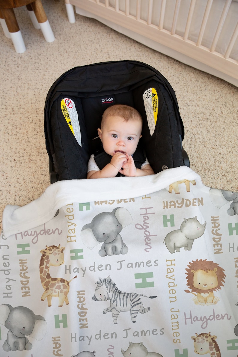 K&ouml;nnte beinhalten: Eine wei&szlig;e Babydecke mit einem sich wiederholenden Muster aus verschiedenen Tieren, darunter eine Giraffe, ein Zebra, ein Elefant, ein L&ouml;we und ein Nashorn. Die Decke tr&auml;gt den Namen "Hayden James" in hellrosa Schrift.