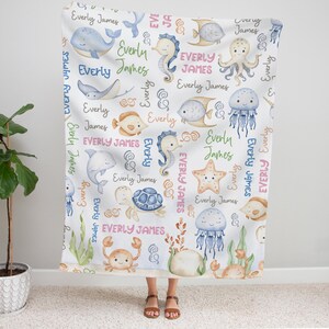 May include: A white baby blanket with a colorful sea creature pattern. The blanket features a variety of sea creatures, including starfish, jellyfish, seahorses, and dolphins. The blanket is decorated with the names "Everly" and "James" repeated throughout the design.