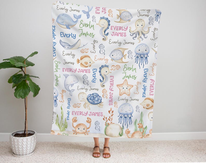 May include: A white baby blanket with a colorful sea creature pattern. The blanket features a variety of sea creatures, including starfish, jellyfish, seahorses, and dolphins. The blanket is decorated with the names "Everly" and "James" repeated throughout the design.