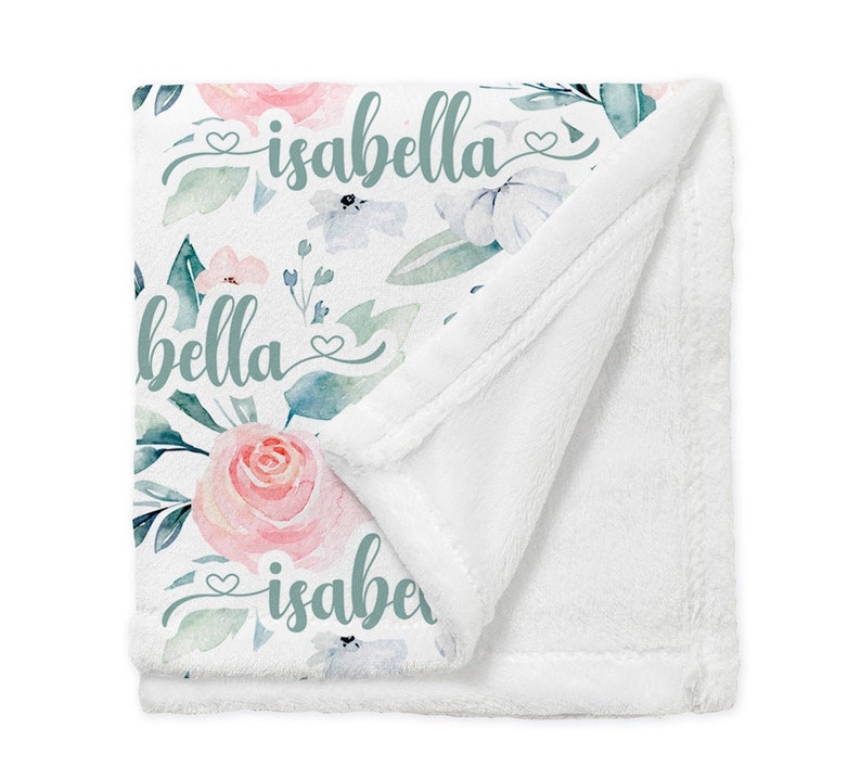 May include: A white baby blanket featuring a floral design and the name "isabella" written in green script. The blanket is made from soft, plush fabric.