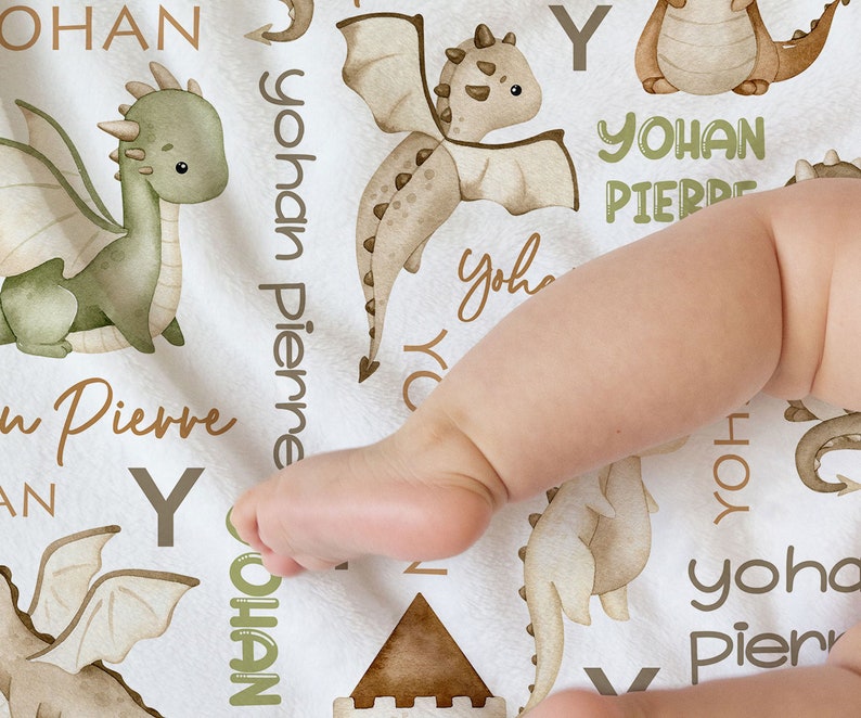 May include: A white baby blanket with a brown and green dragon design. The blanket features the names "Yohan Pierre" and "Yohan" repeated throughout the design. The blanket is made of soft, comfortable fabric and is perfect for a baby's nursery.