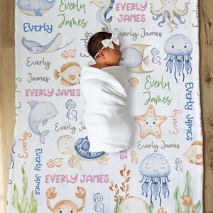 May include: A white baby blanket with a sea life theme. The blanket features a variety of sea creatures, including a whale, a seahorse, an octopus, a jellyfish, a starfish, a turtle, and a crab. The blanket is decorated with the name "Everly James" repeated throughout.